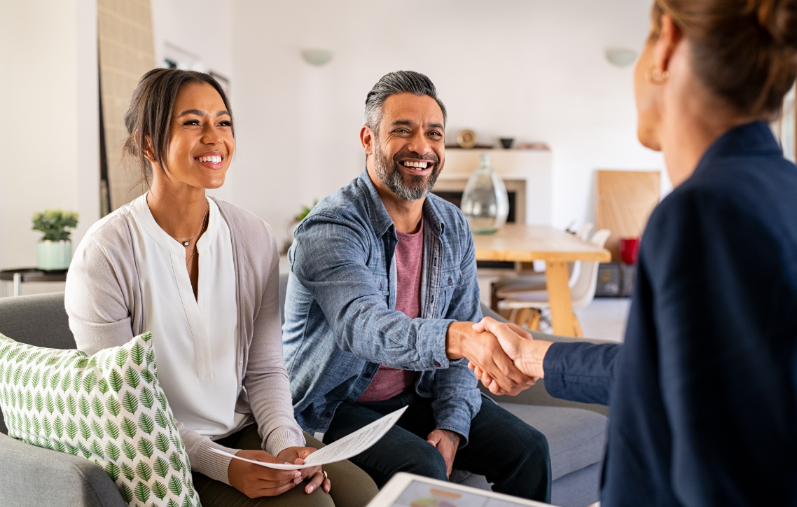 Two people smiling and shaking hands in a bright living room during a meeting
