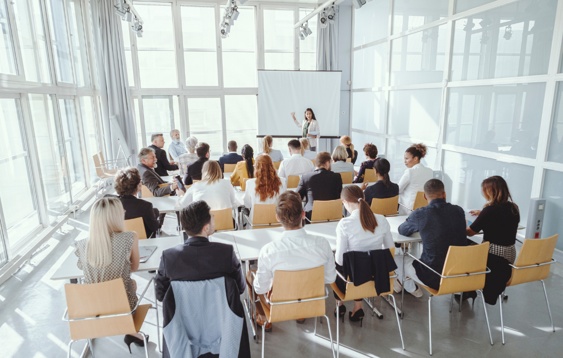 People seated around a conference table in a bright modern meeting room, listening to a presenter.