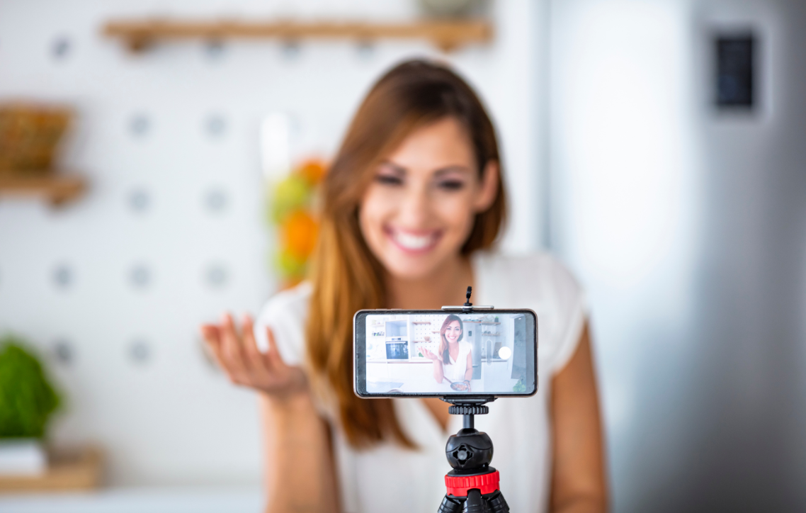 Woman filming a selfie video on a smartphone mounted on a small tripod, smiling in a bright room