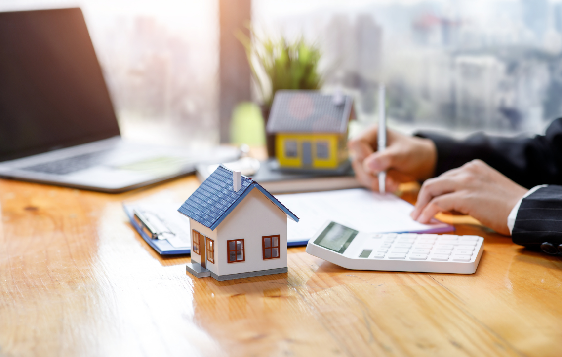 Model house on a desk beside a person reviewing papers and a calculator in a bright office