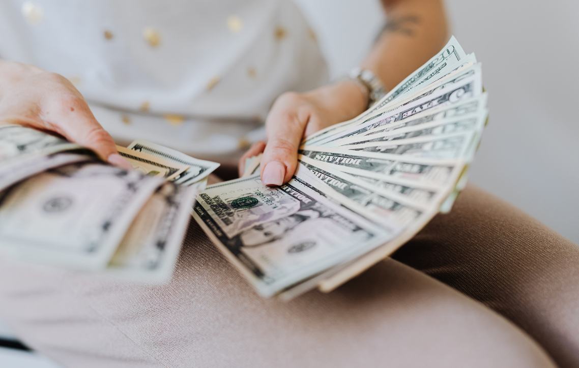 Hands fanning U.S. dollar bills over a beige surface, with more cash in the foreground