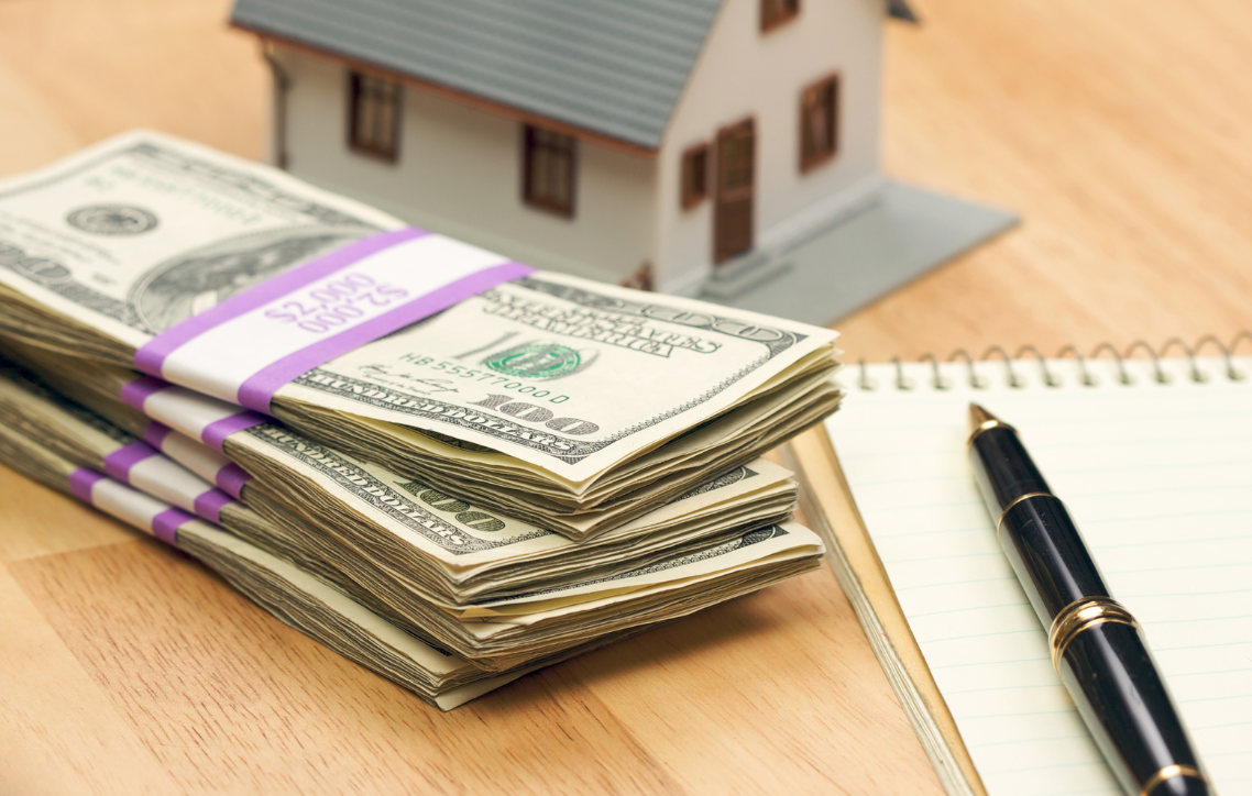 Stack of cash tied with purple bands beside a model house, notebook, and pen on a desk.