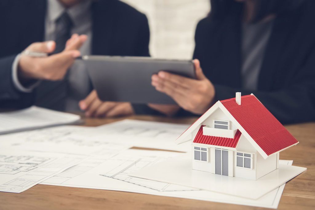 A man and woman are looking at a tablet while a model house sits on a table.