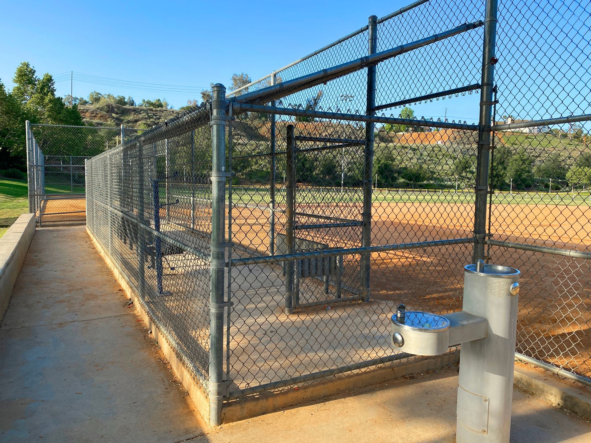 Dog park enclosure with benches and water fountain, next to a path and dirt field.
