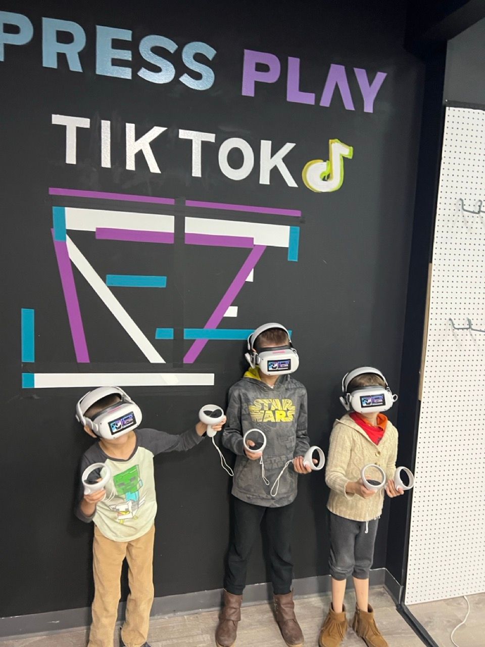 Three children wearing virtual reality headsets in front of a wall that says press play tik tok