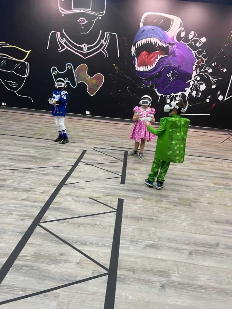 Three children in costumes are playing in a room with a dinosaur on the wall behind them