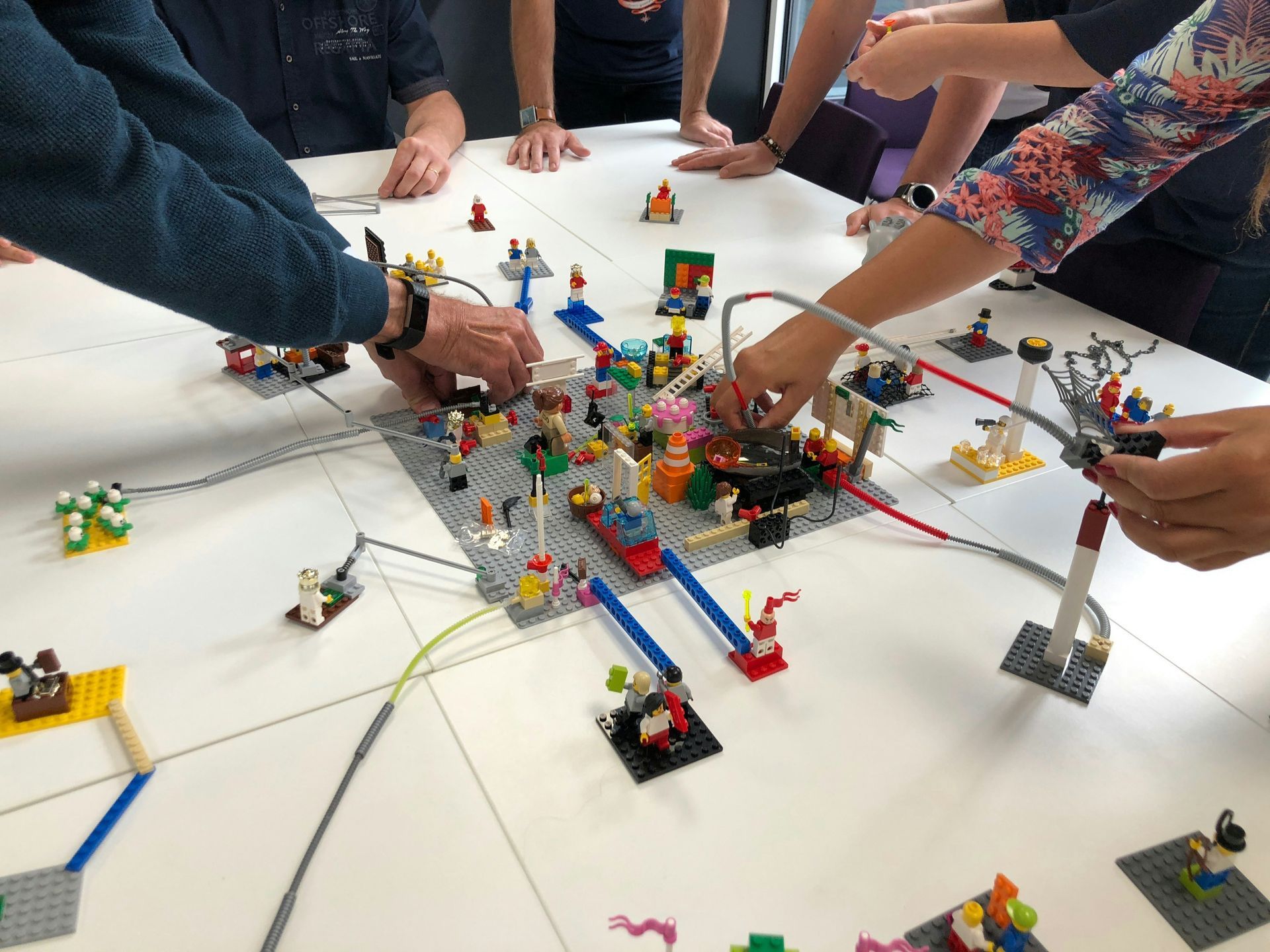 Hands reaching to modify a Lego city on a white table. Several people gather around, wires connecting different structures.