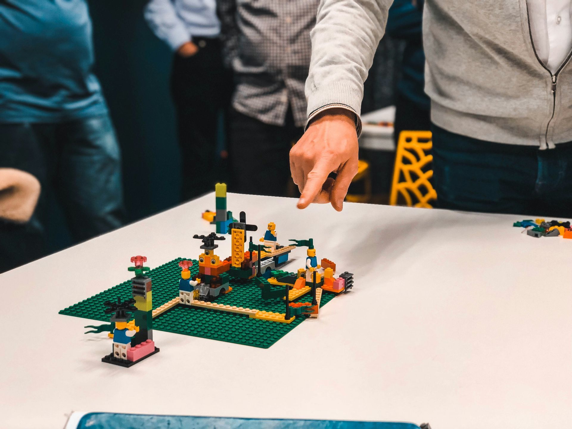 Person points to LEGO structure on a white table; other people and LEGO bricks are also present.