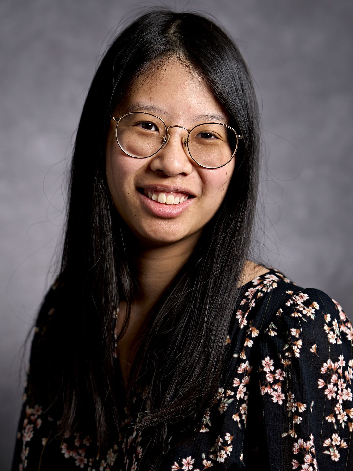 A woman with long black hair is wearing a floral shirt and smiling.