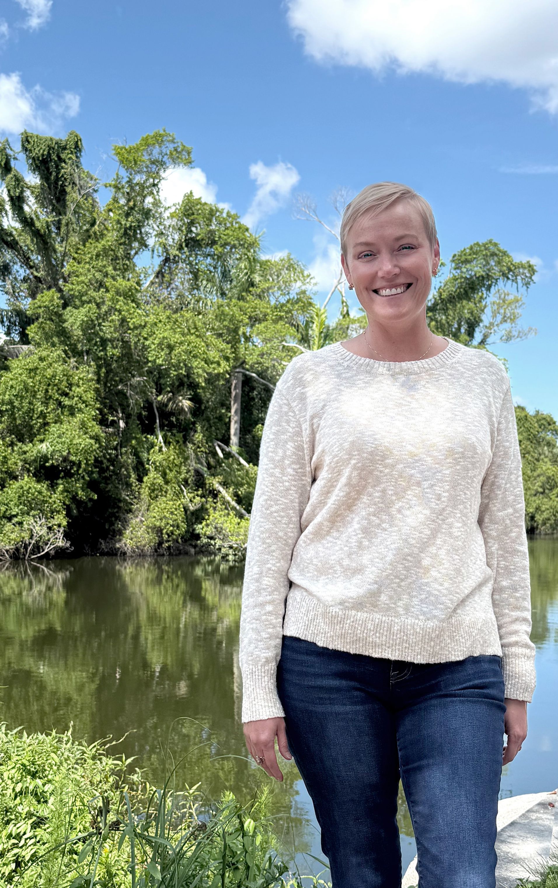 A woman in a white sweater and jeans is standing in front of a river.