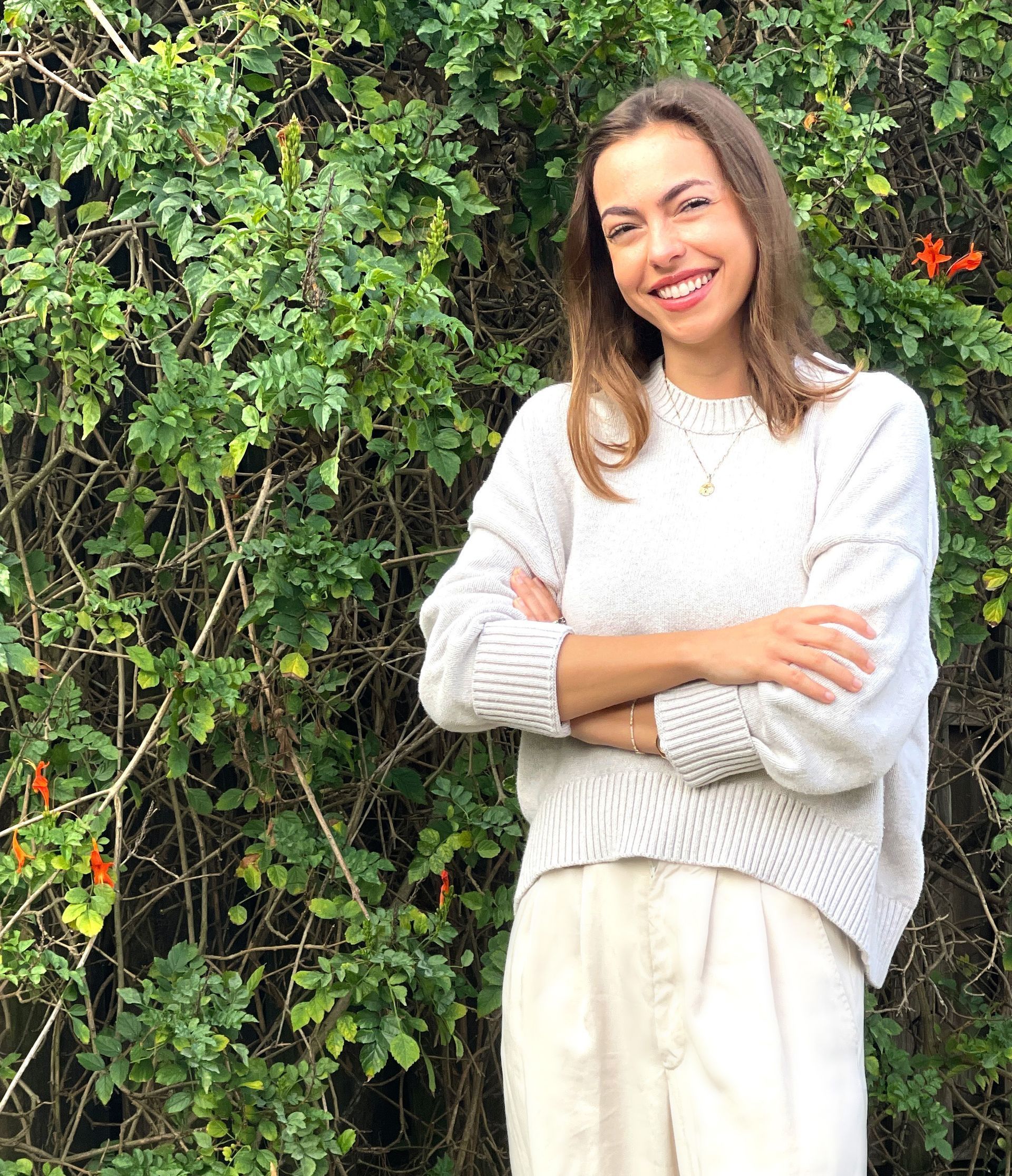 A woman in a white sweater and white pants is standing in front of a bush with her arms crossed.