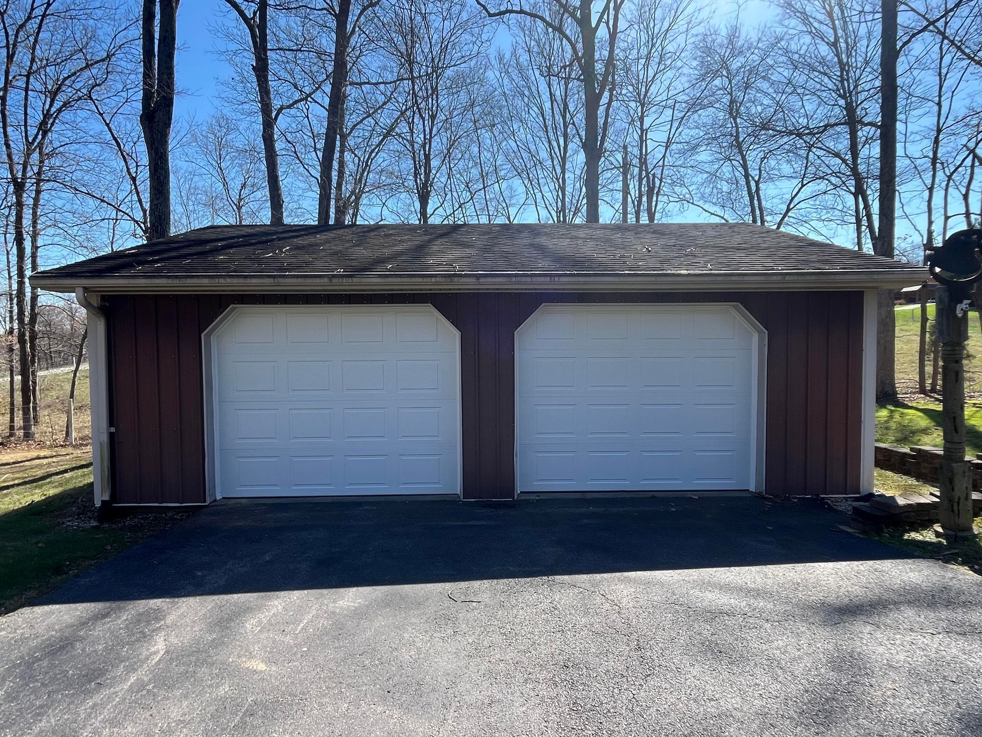 A garage with two white garage doors is sitting in the middle of a forest.