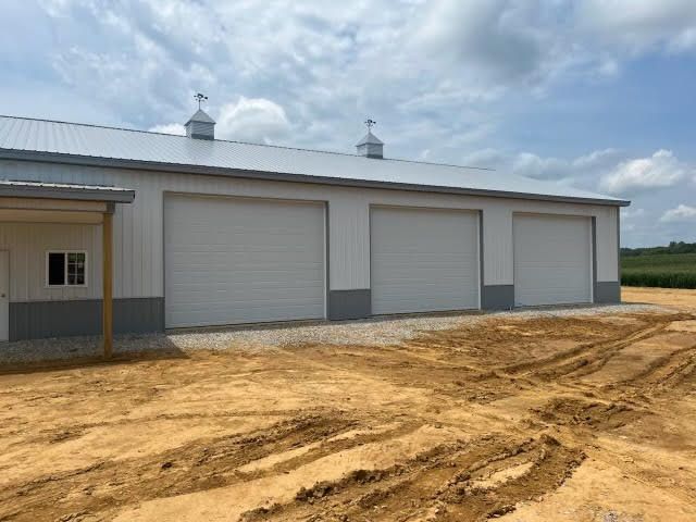 A large white building with three garage doors is sitting in the middle of a dirt field.