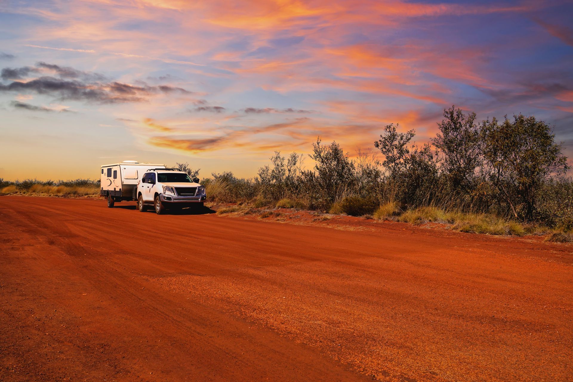 A white SUV towing a travel trailer parked on a red dirt road under a vibrant sunset sky in an arid landscape.