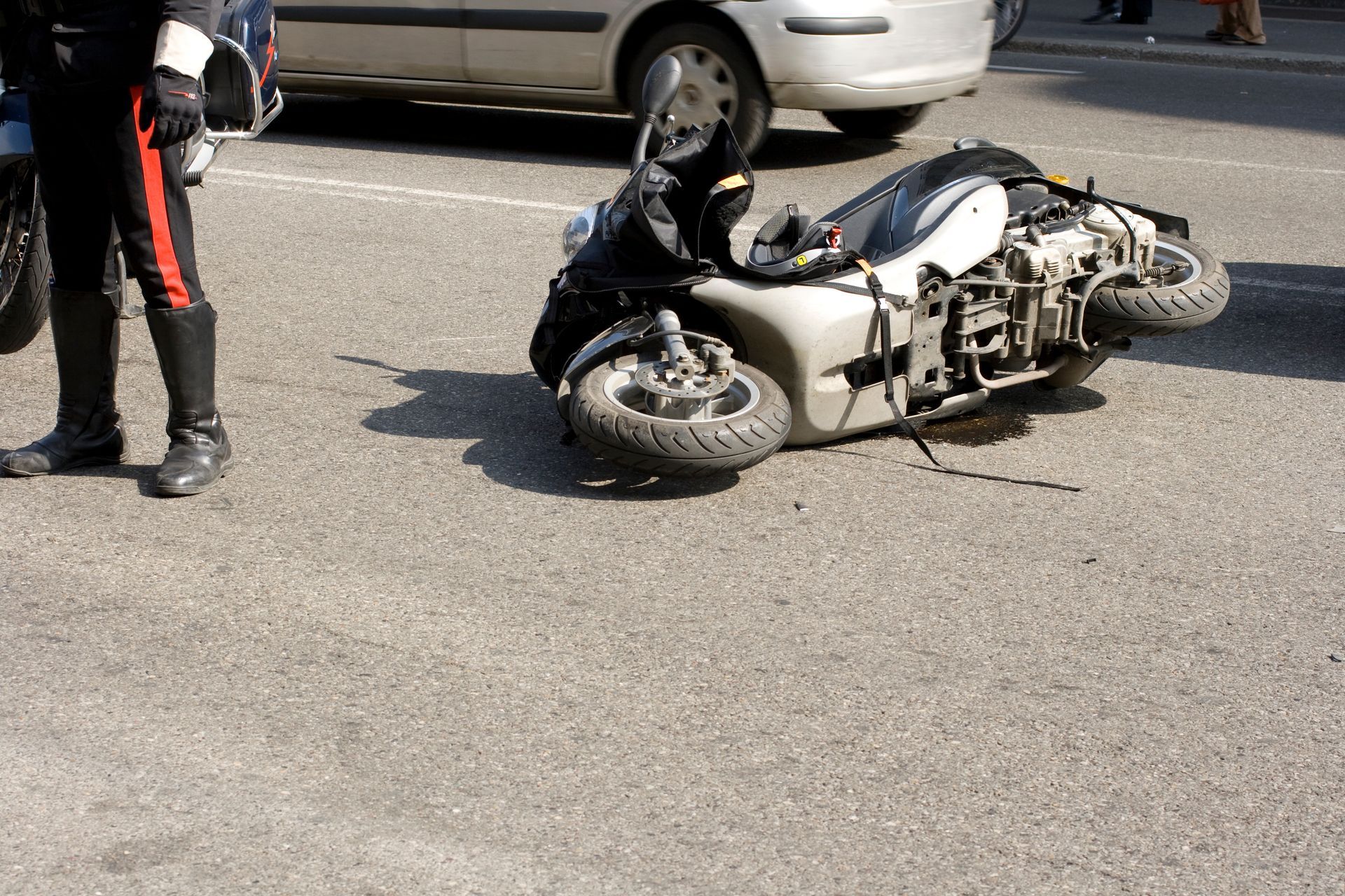 A white and black scooter lying on its side on an asphalt road next to the legs of a uniformed person.