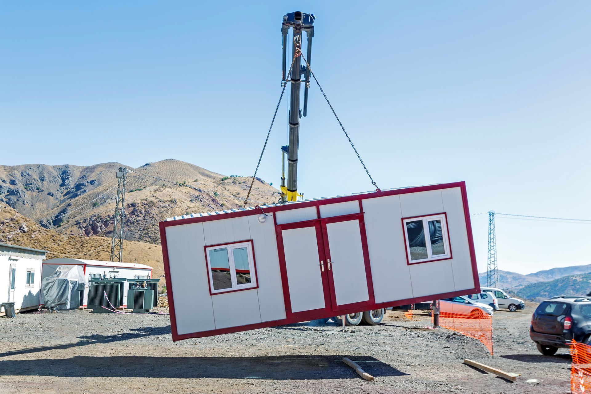 A crane lifts a white modular building unit with red trim against a clear blue sky and arid mountain landscape.