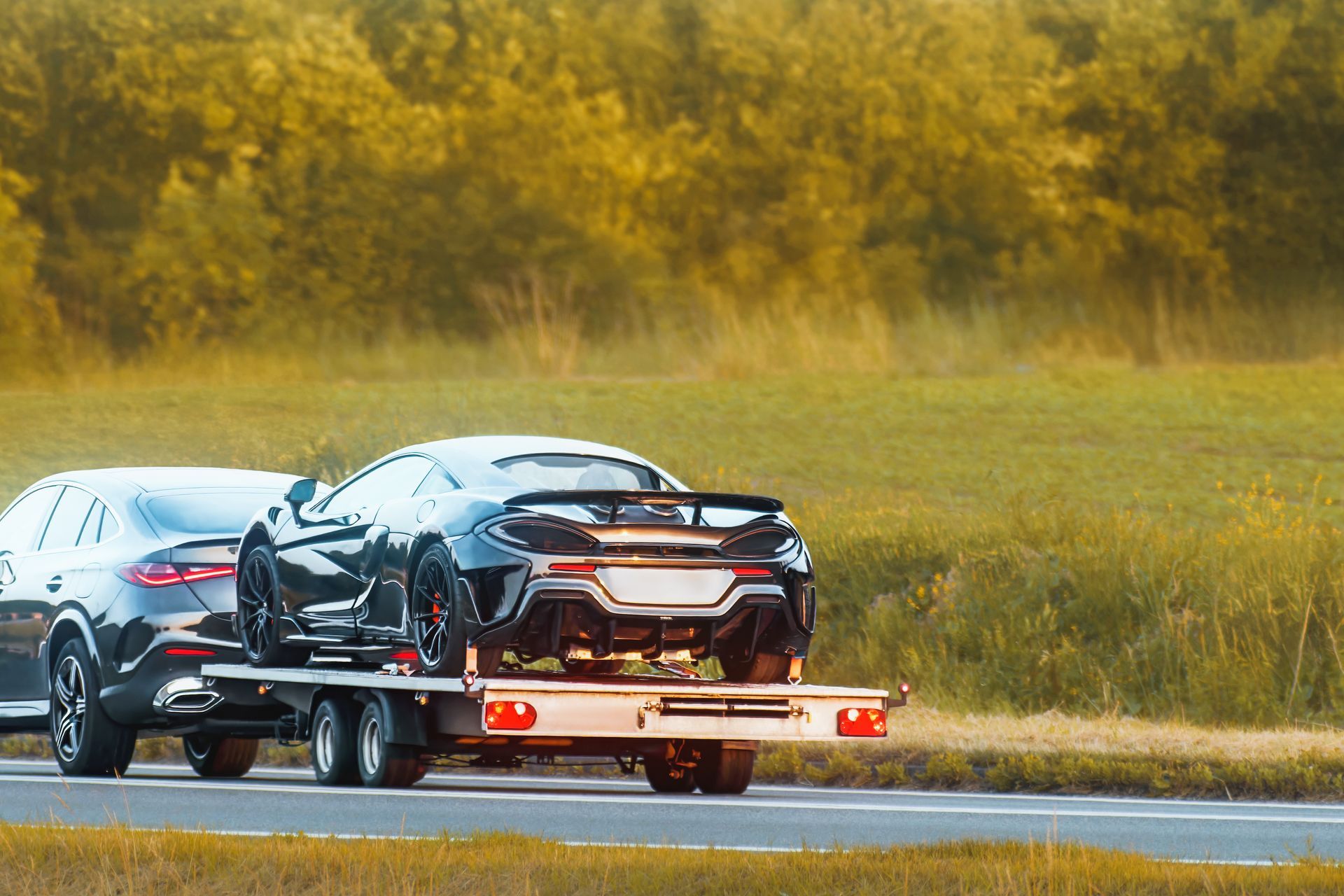 A black McLaren supercar on a car trailer being towed by a vehicle along a road near a green field.