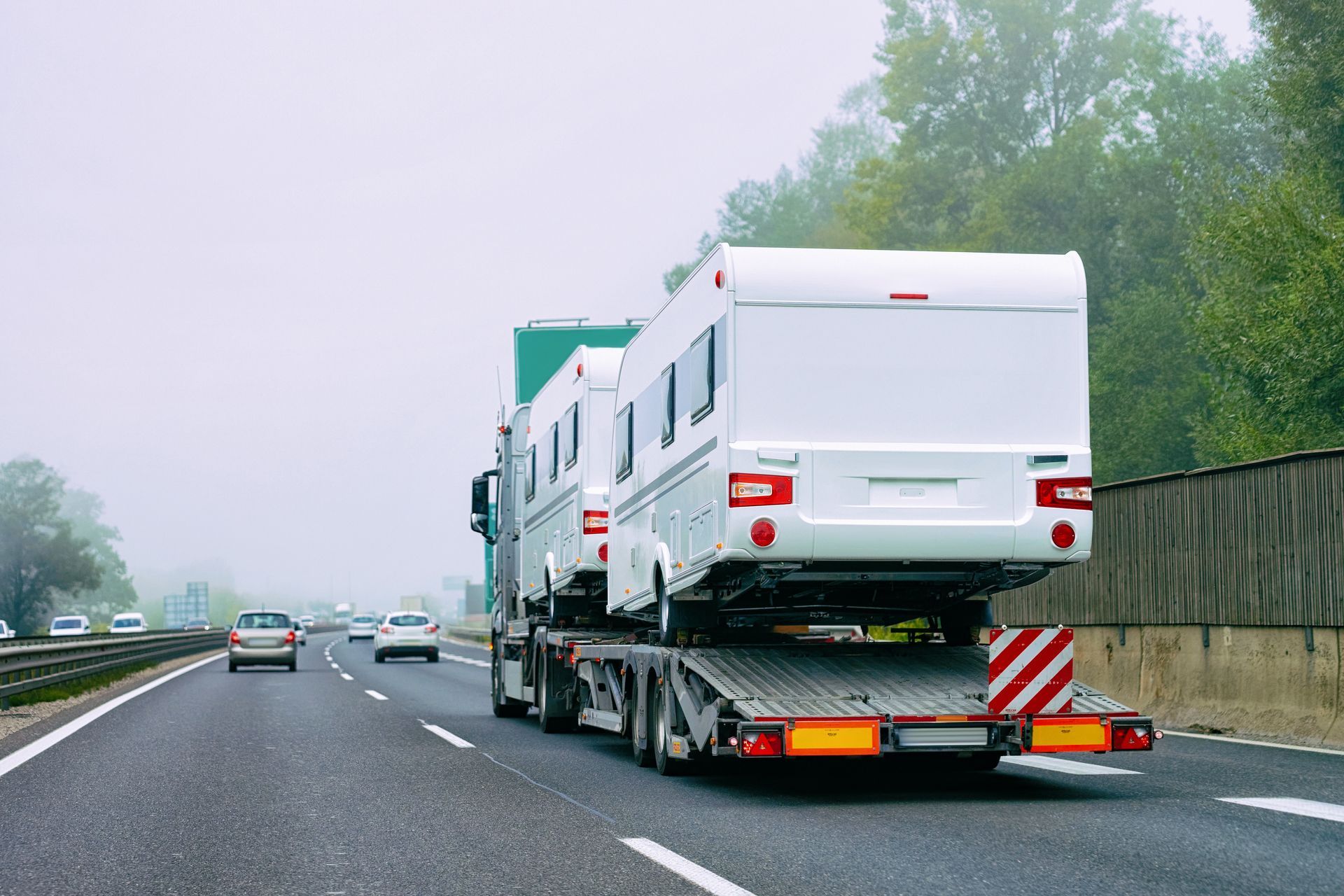 A car hauler truck on a highway carrying two white travel trailers in foggy conditions.
