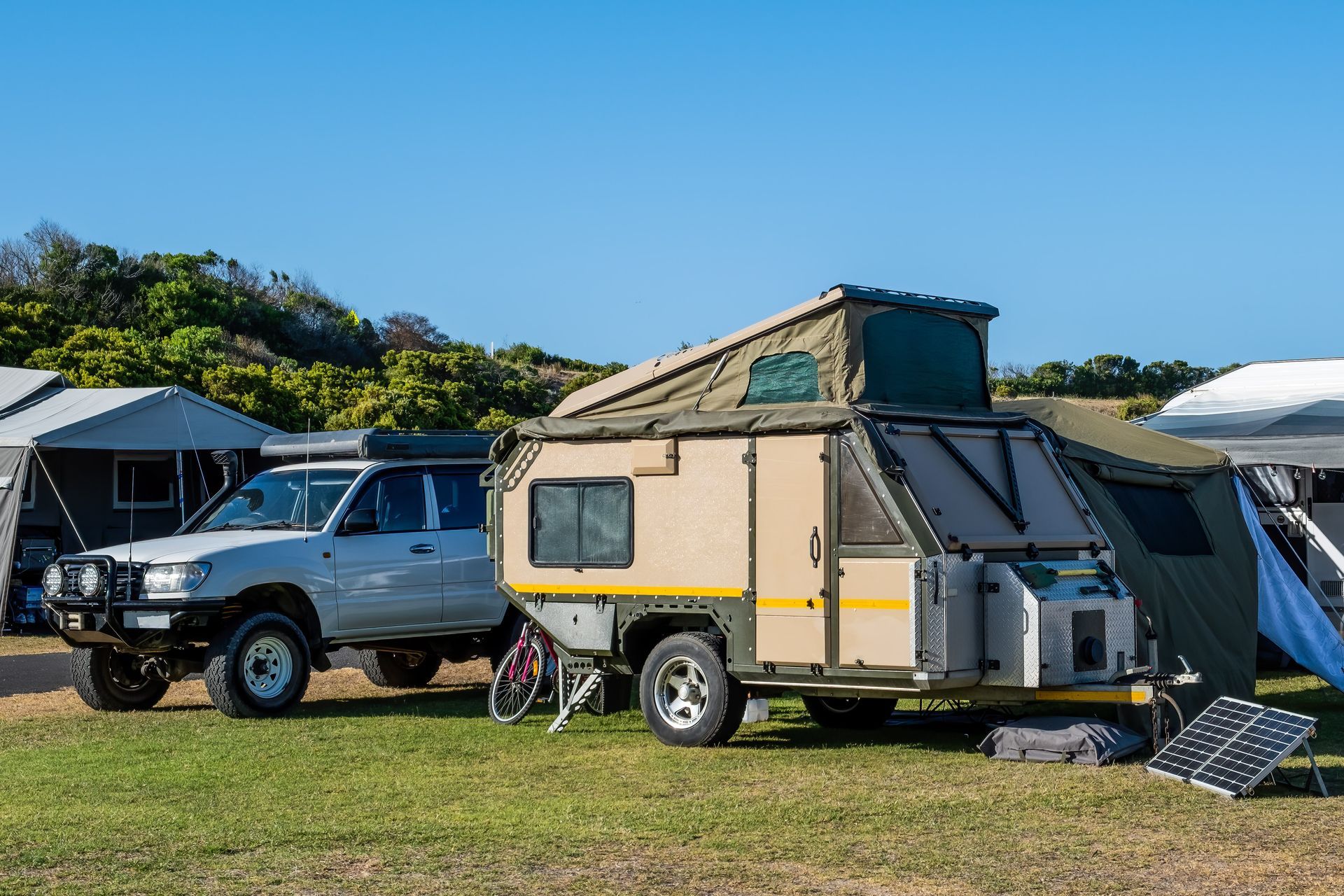 A silver 4x4 vehicle parked on a grassy field, towing a tan pop-up camper with a solar panel on the ground nearby.
