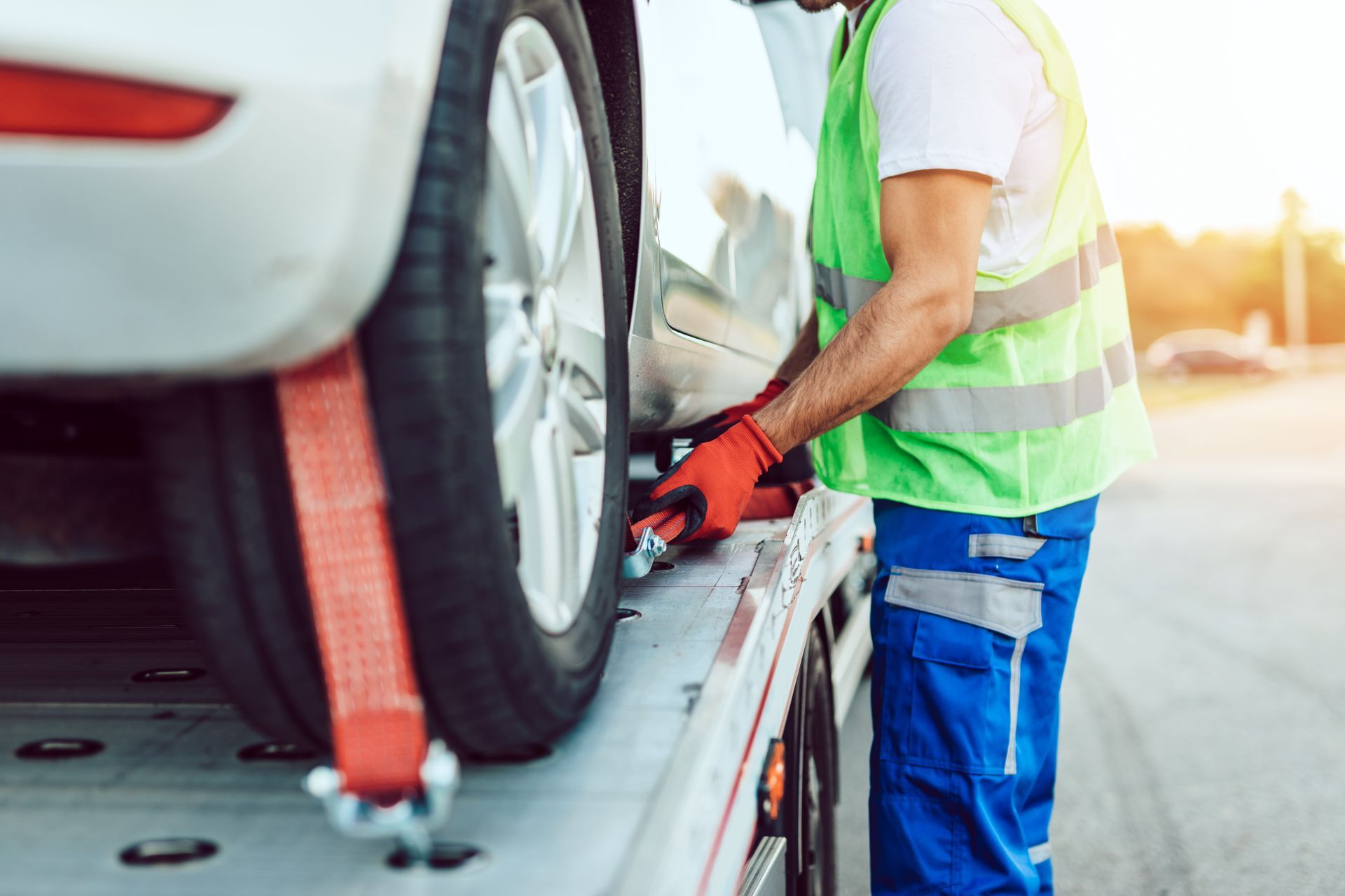 A worker in a high-visibility vest secures a vehicle to a flatbed tow truck with an orange strap.