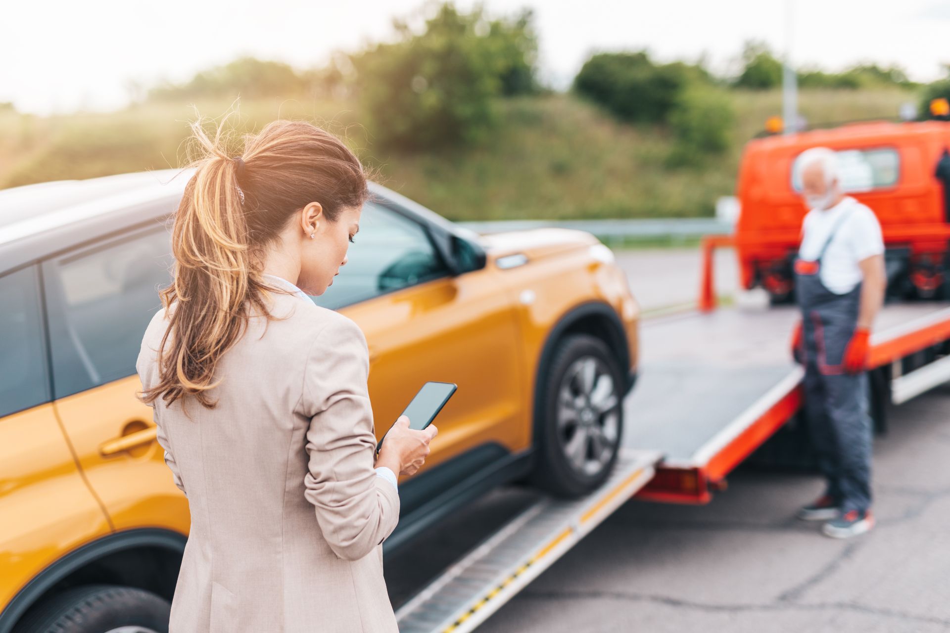 A woman looks at her phone while a tow truck driver loads her yellow car onto a flatbed trailer outdoors.