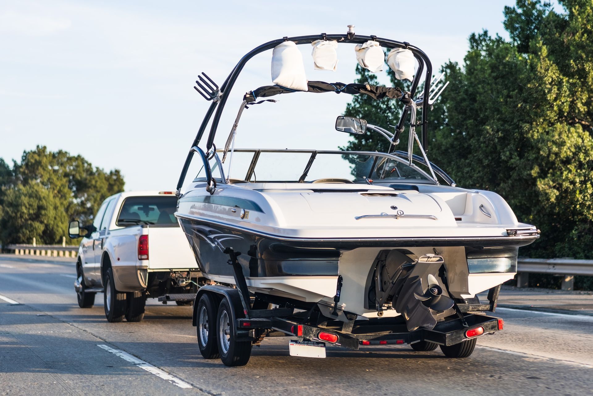 A white truck towing a black and white motorboat with a black wakeboard tower on a highway surrounded by trees.