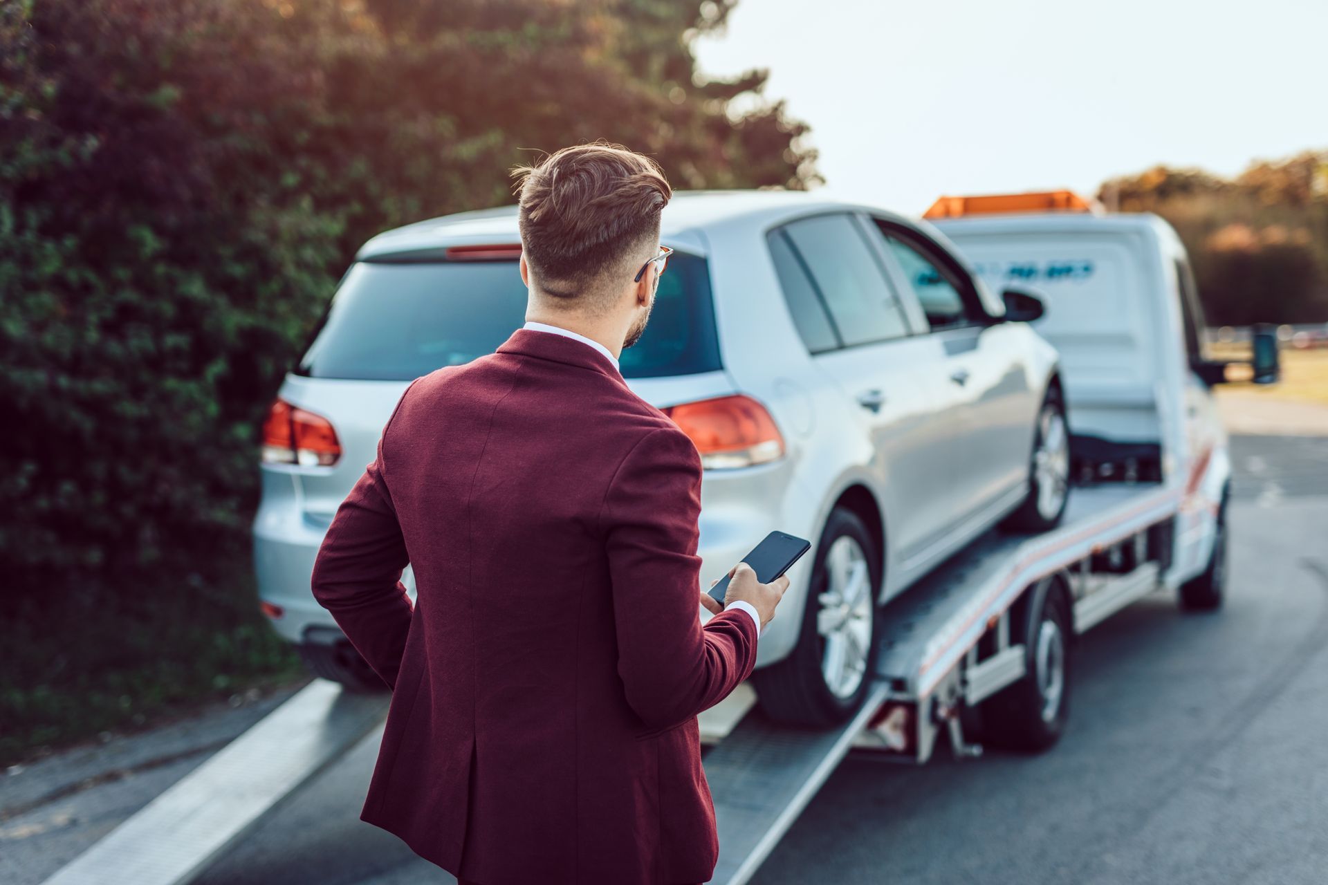 A person in a maroon blazer holds a smartphone while a silver car is being loaded onto a tow truck.