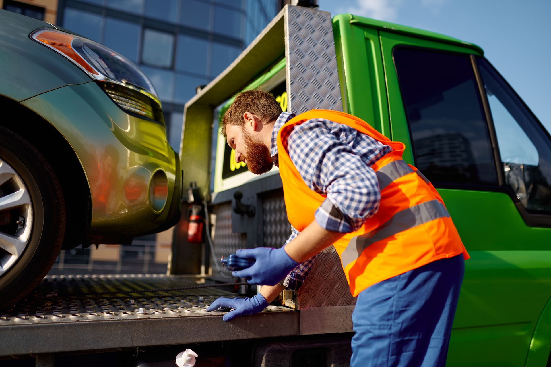 A worker in an orange safety vest and blue gloves secures a green car onto the bed of a green tow truck.