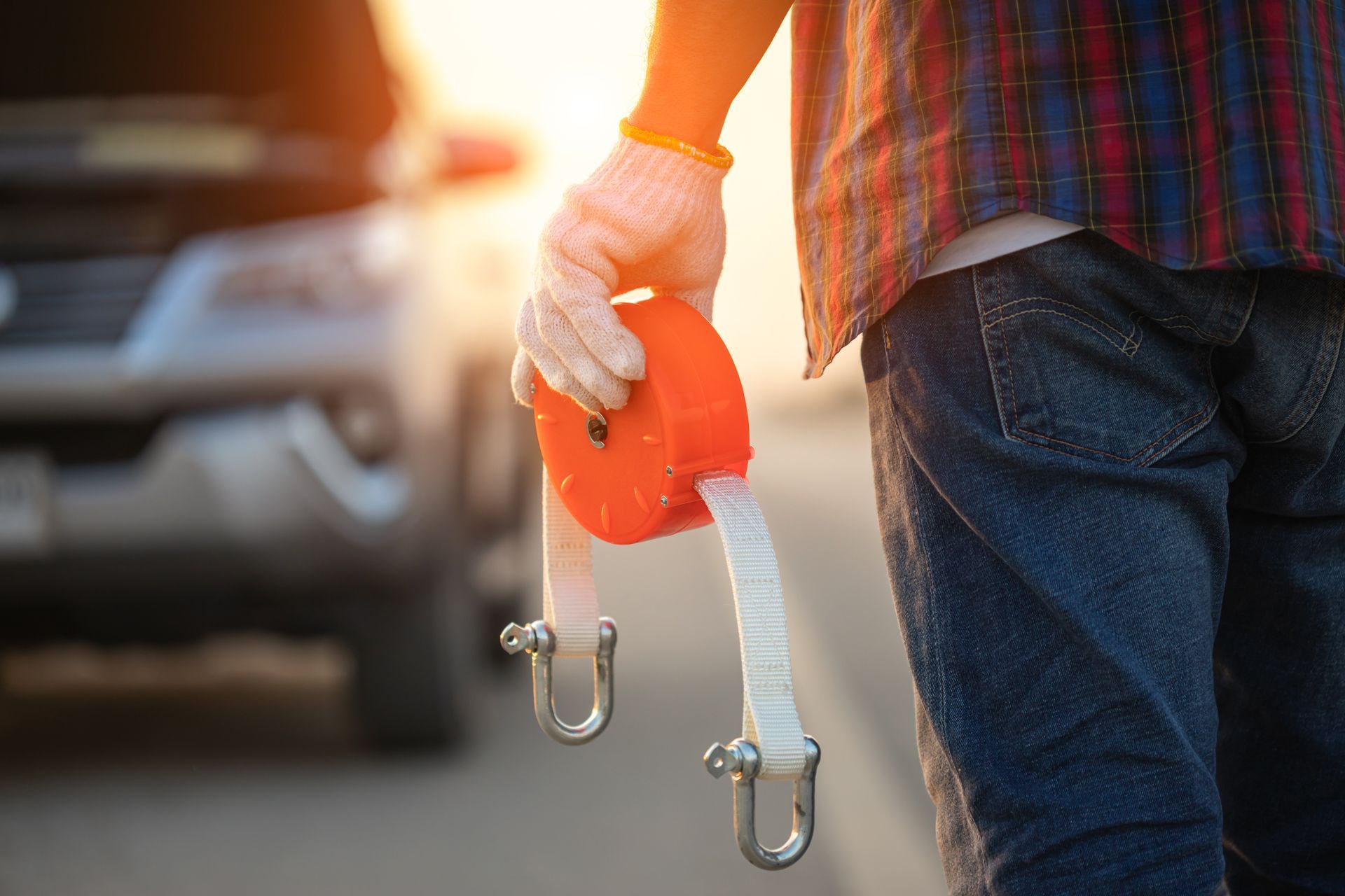 A person holds an orange tow strap with metal shackles in front of a car with its hood raised on a road at sunset.