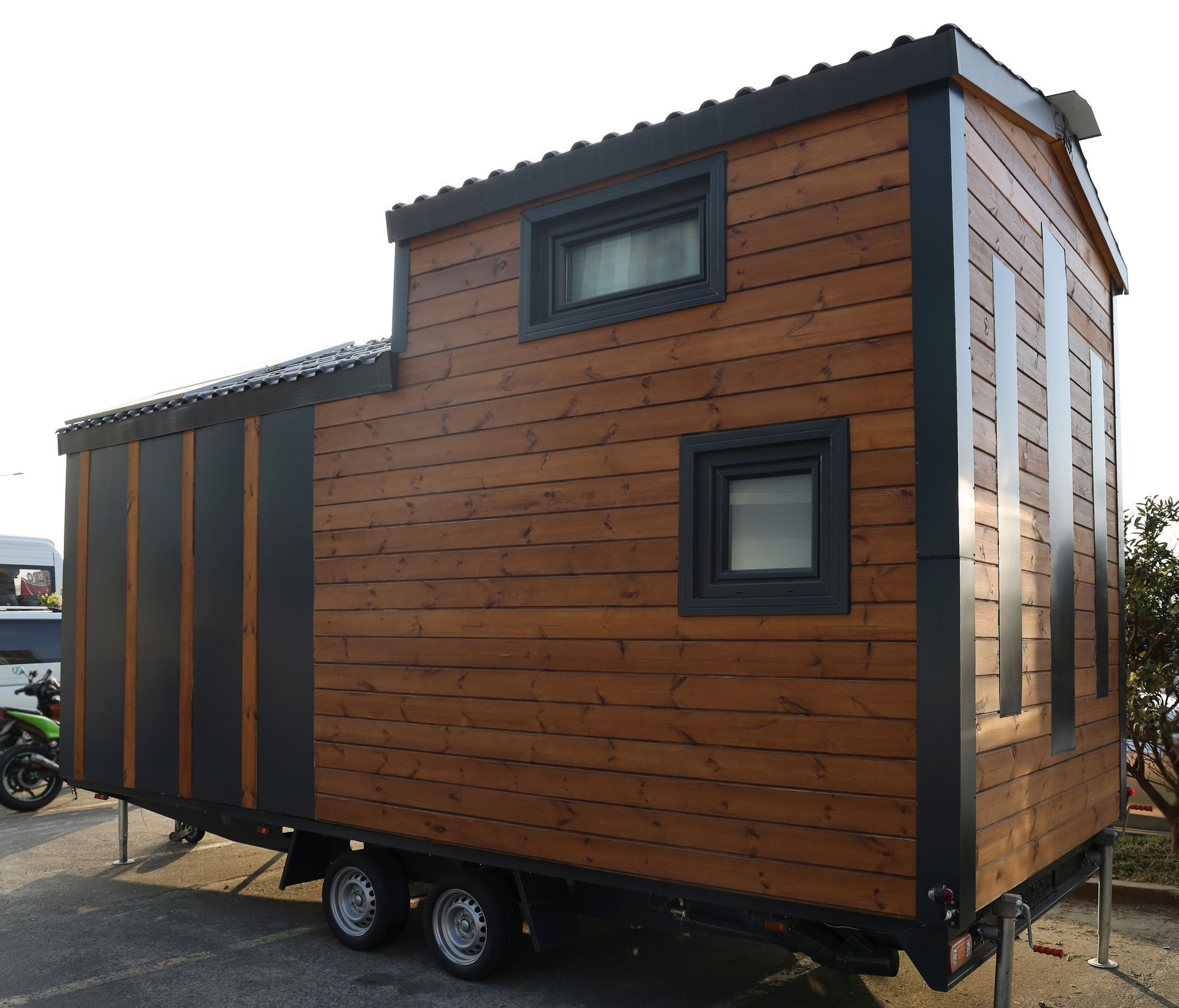 A modern tiny house on a trailer with wood siding and dark trim, featuring a loft window and a ground-level window.
