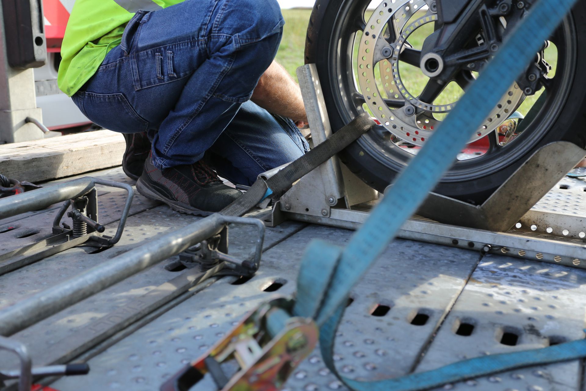 A person in a neon shirt and jeans secures a motorcycle wheel to a flatbed trailer using blue ratchet straps.