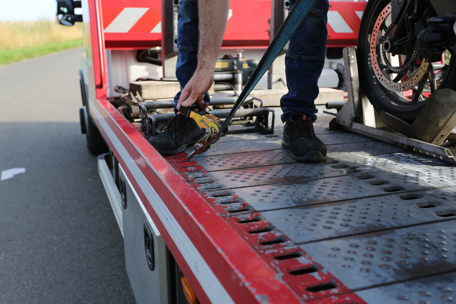 A person secures a motorcycle to the bed of a red flatbed tow truck on the side of a road.