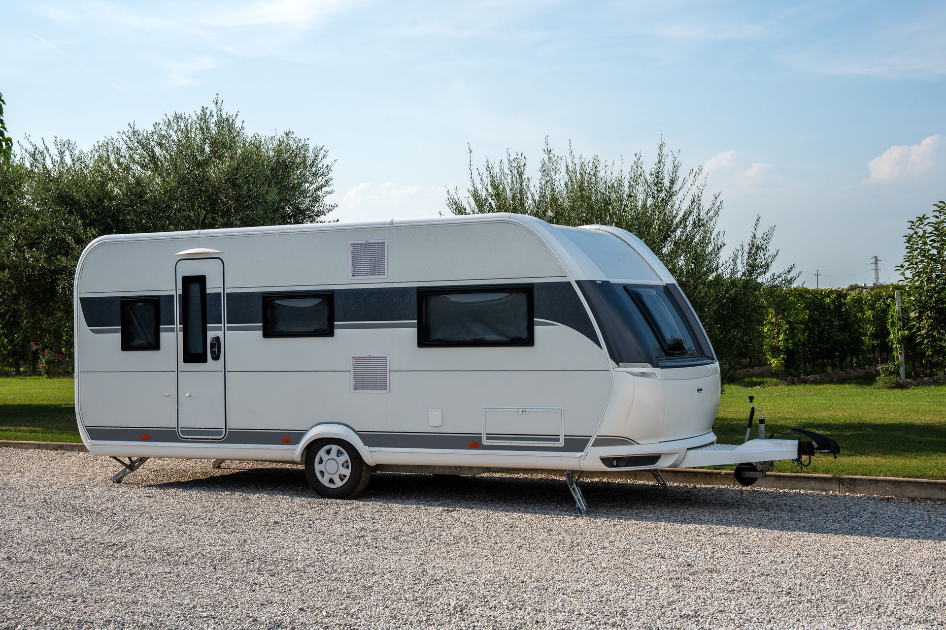 A white travel trailer with grey detailing parked on a gravel surface against a blue sky and green trees.