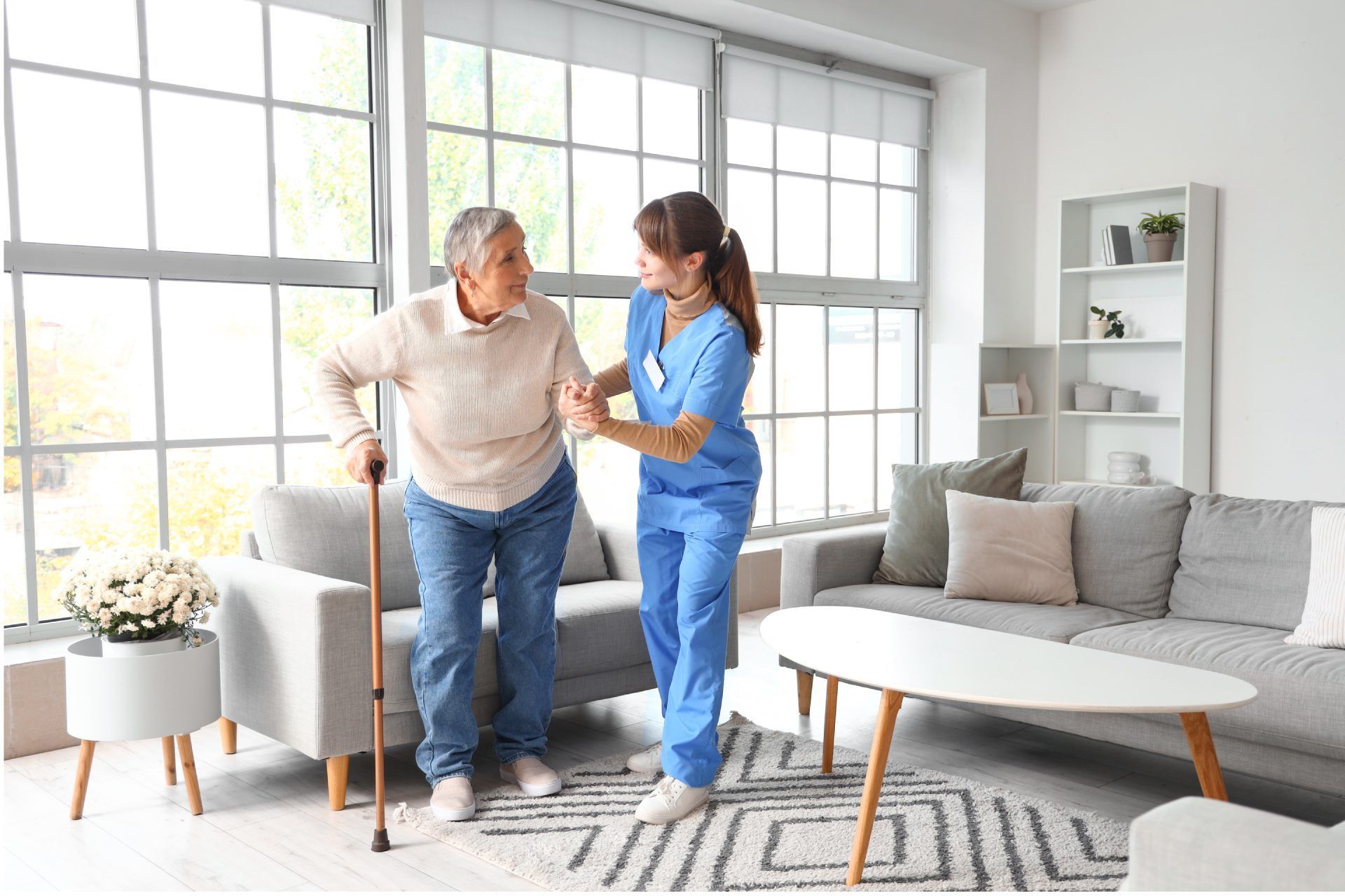 Nurse assists elderly woman with a cane to walk in a living room with natural light.