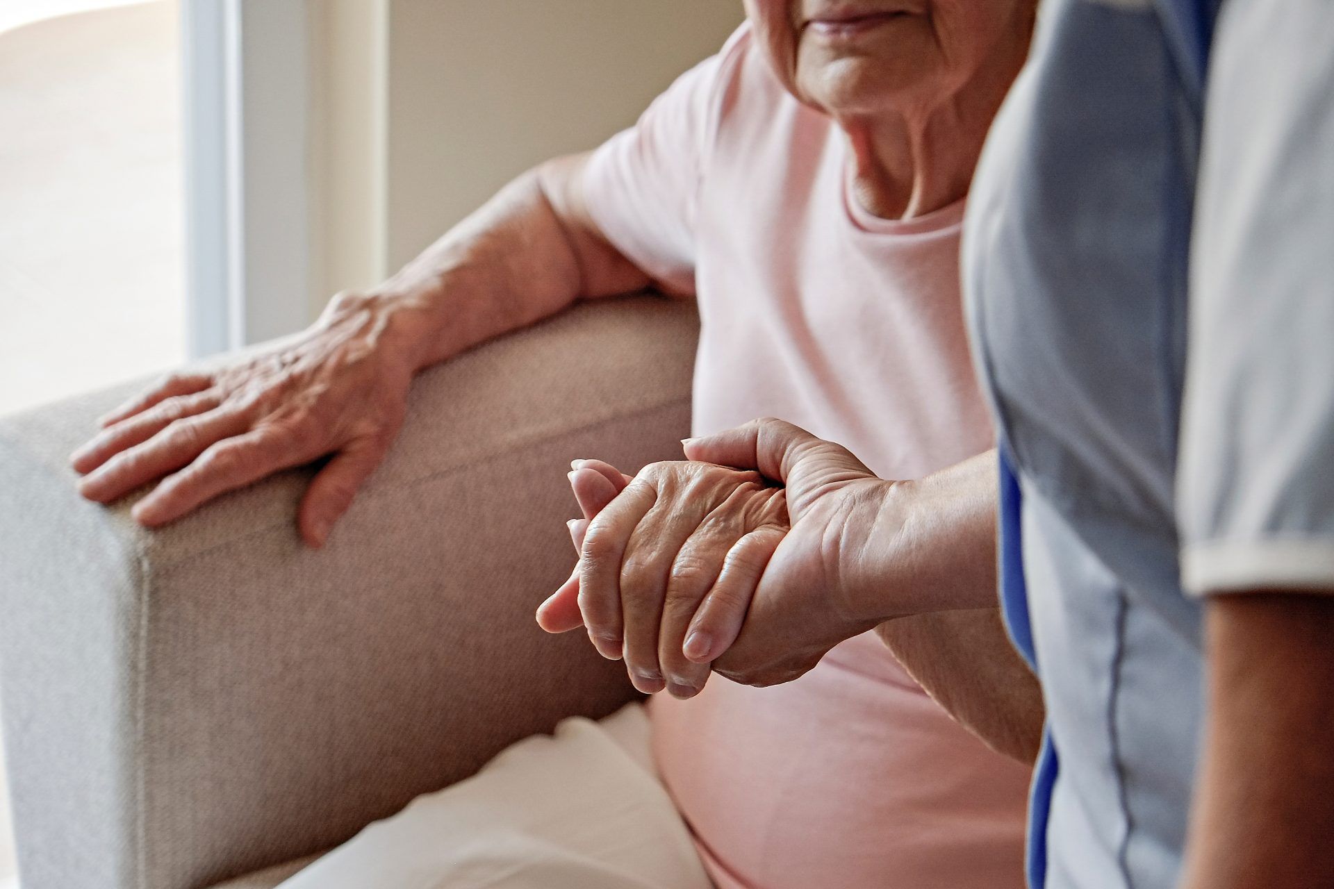 A caregiver holds the hand of an elderly woman, offering support in a light-filled room.