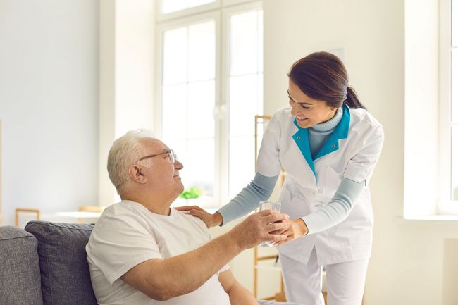 Caregiver assisting elderly man, indoors. She holds a glass of water, smiling, while he sits on a couch.