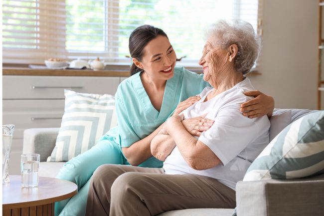 Caregiver embracing and smiling with elderly woman on a couch in a bright living room.