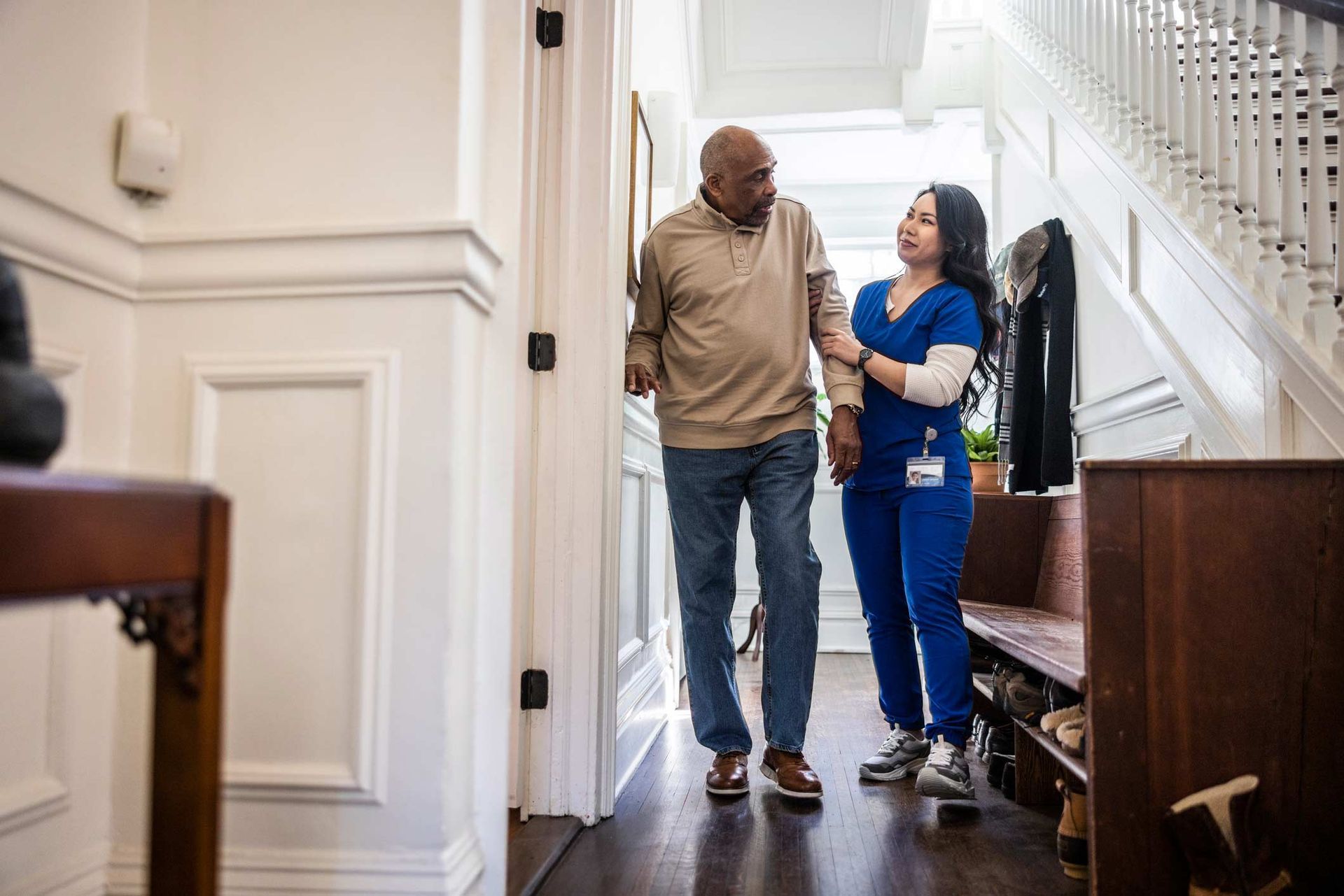 A woman in blue scrubs assists an elderly Black man walking down a hallway with stairs.