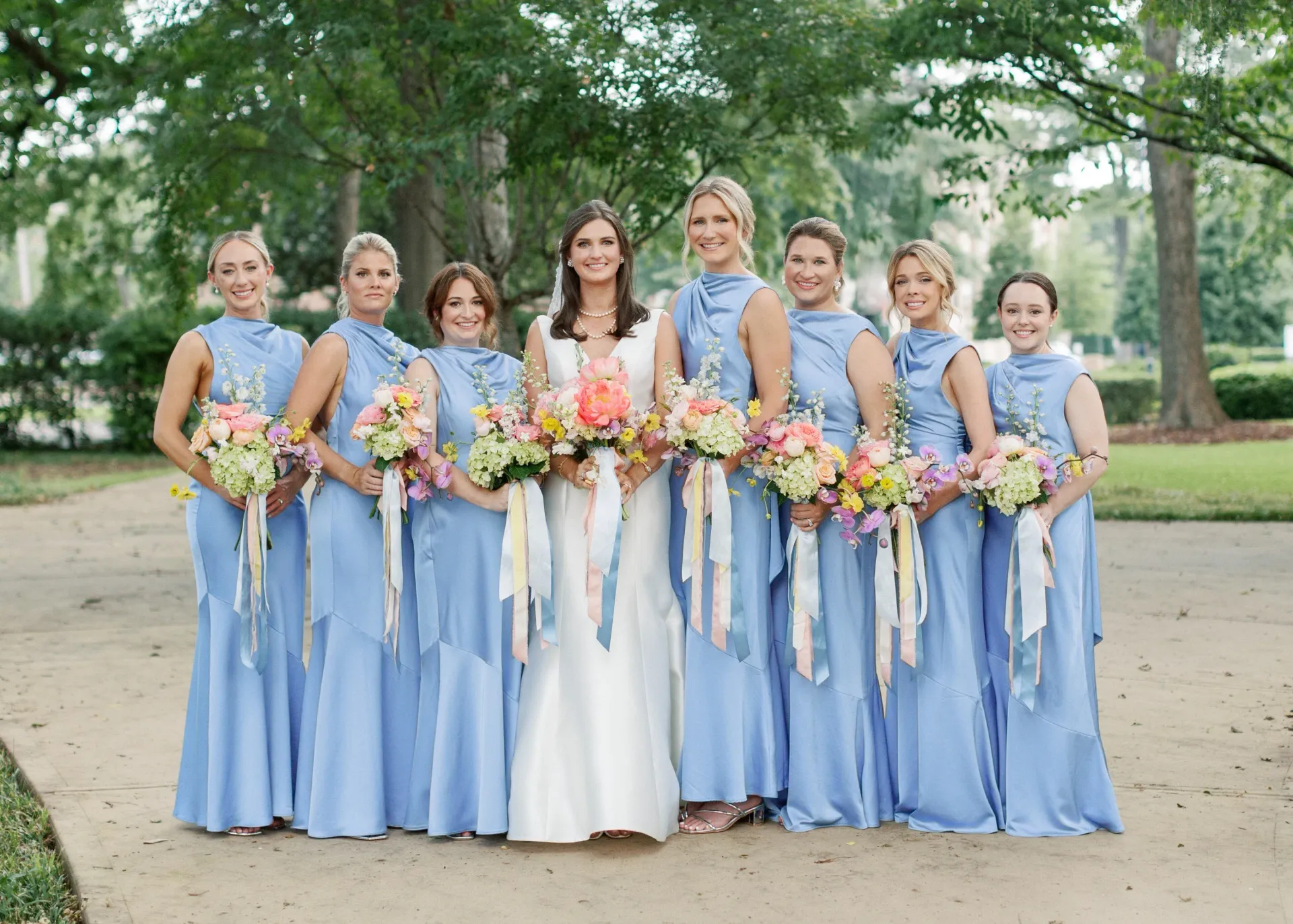 Bride and bridesmaids in blue dresses, holding bouquets outdoors.