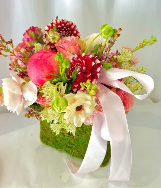 Floral arrangement in moss-covered square container, with pink and red flowers, tied with a white ribbon.