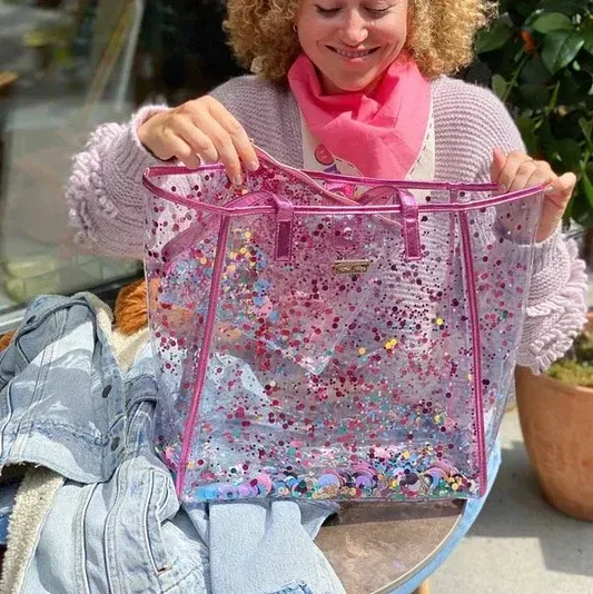 Woman holding a clear pink tote bag filled with confetti, smiling, with denim jacket nearby.