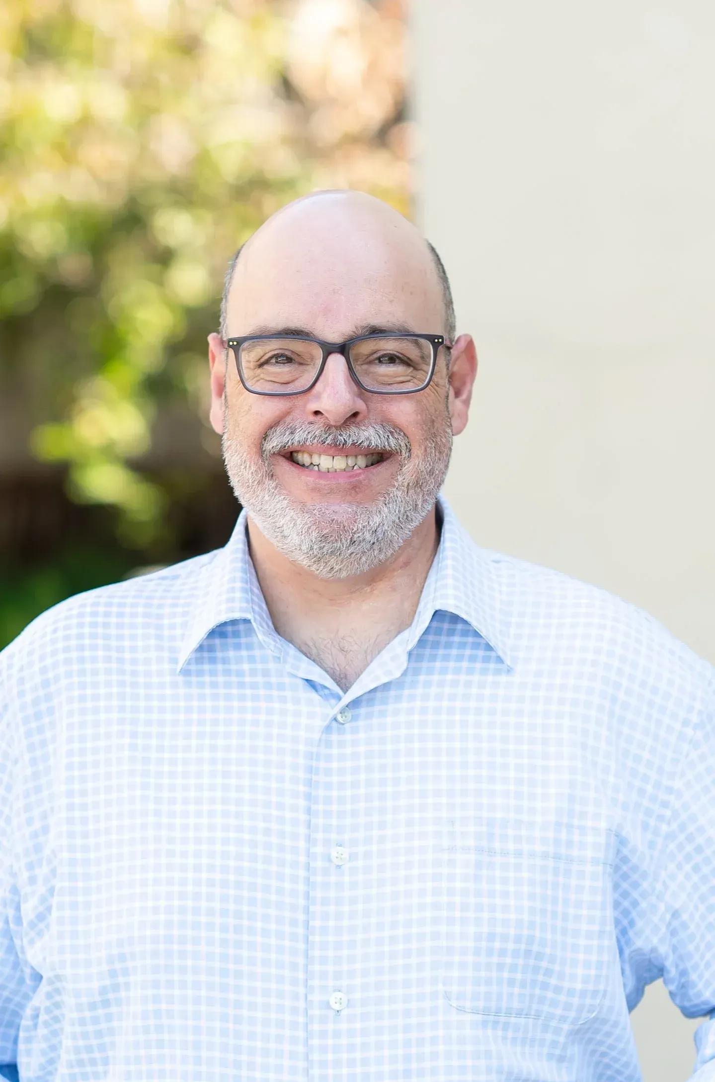 Man with glasses, bald head, and beard smiling, wearing a light blue checkered shirt, outdoors.