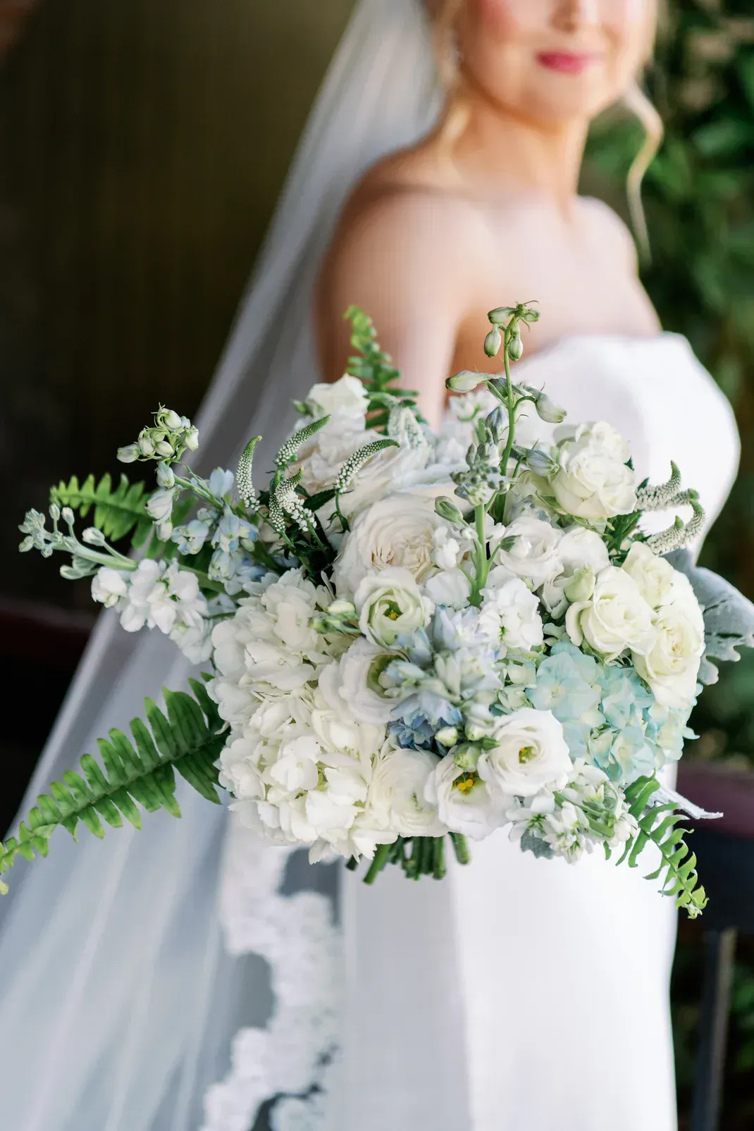 Bride in white dress holding a bouquet of white and blue flowers, with a veil.