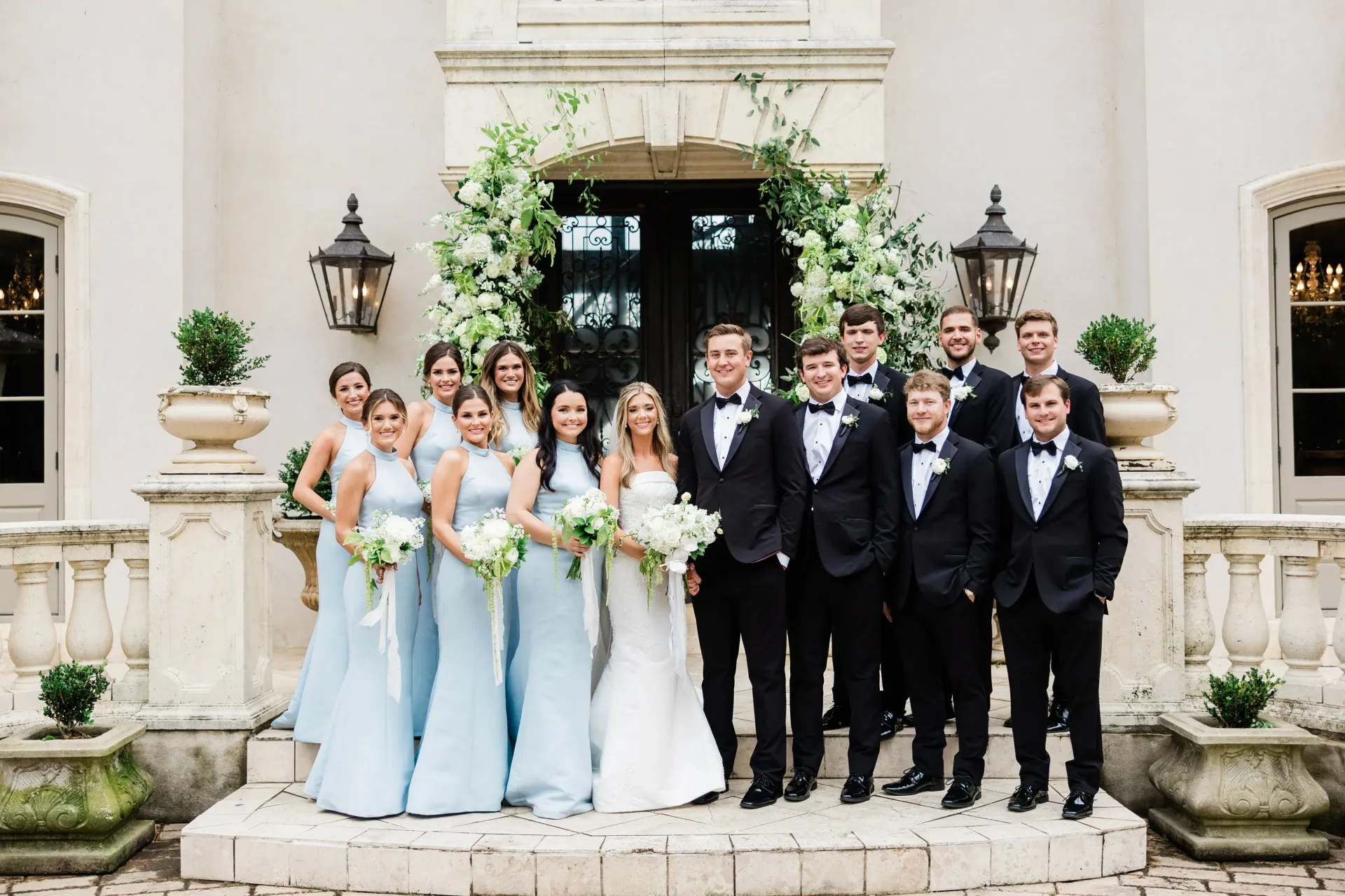 Wedding party posing in front of a white building with floral archway. Bride and groom centered; bridesmaids in blue, groomsmen in black.