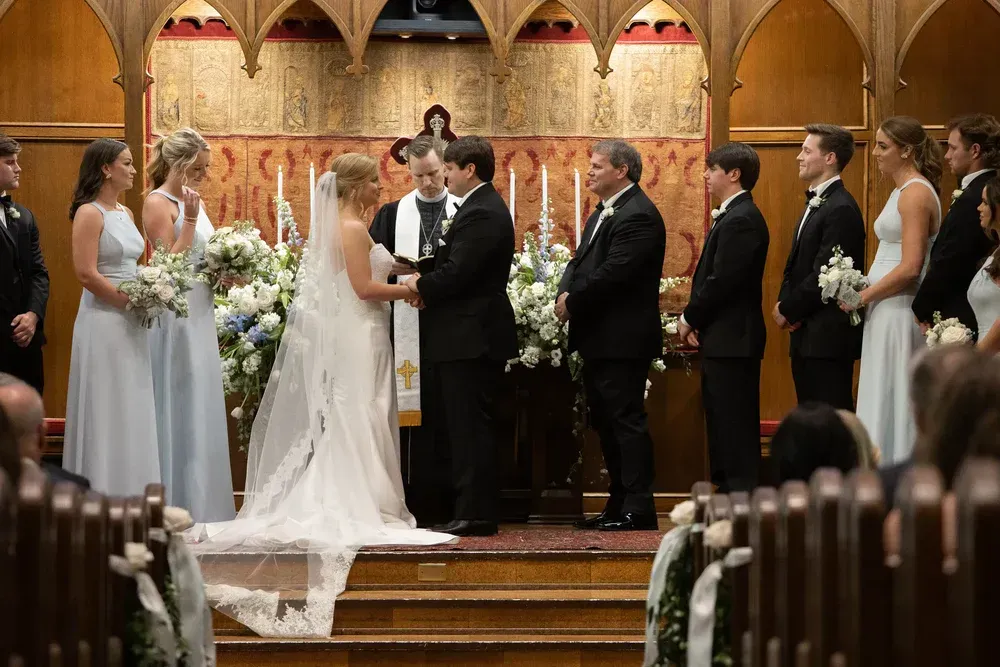 Wedding ceremony in a church. Bride and groom exchange vows, flanked by wedding party and officiant.