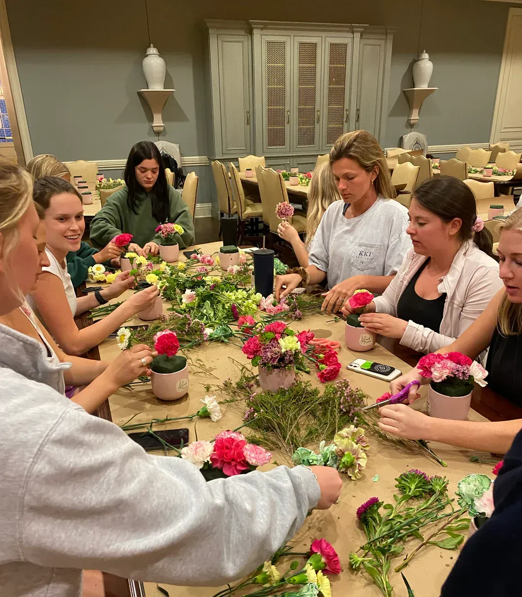 People arranging flowers at a table; various pink and green blooms and small pots, indoor setting.