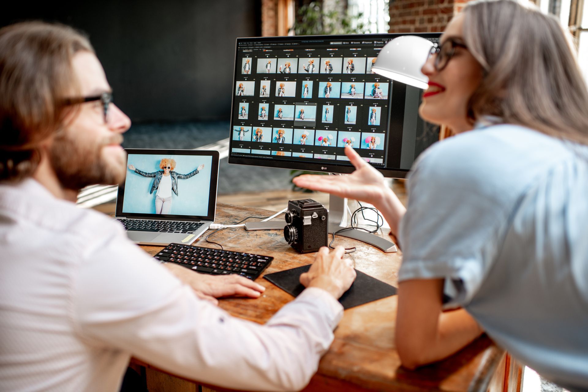 Two people reviewing photos on a computer and laptop at a desk. The woman gestures, smiling.