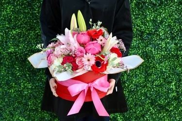 Person holding a red hatbox overflowing with pink and red flowers, tied with a pink bow, against a green background.