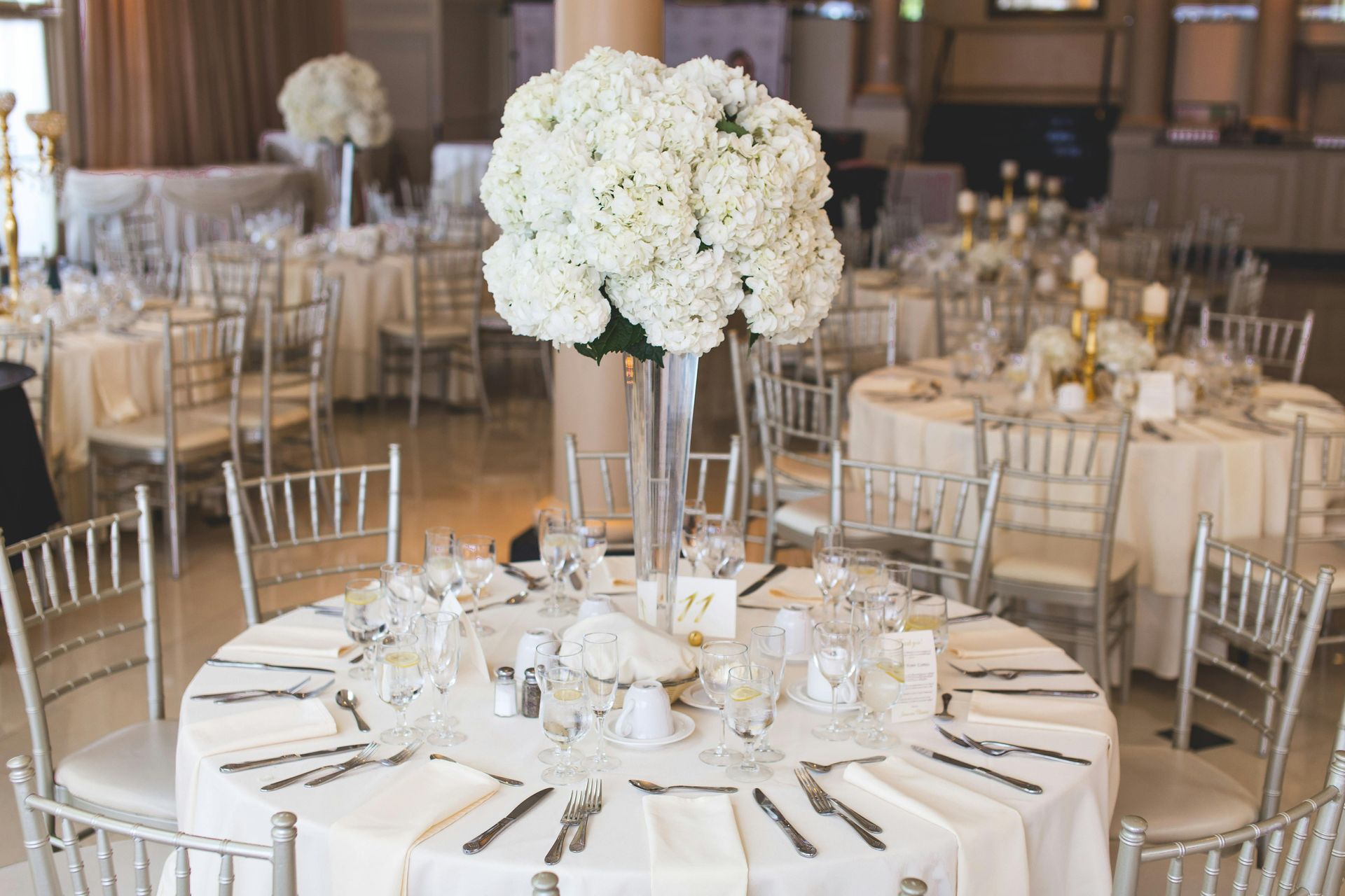 Elegant wedding reception with round tables, white linens, silver chairs, and large white floral centerpieces.