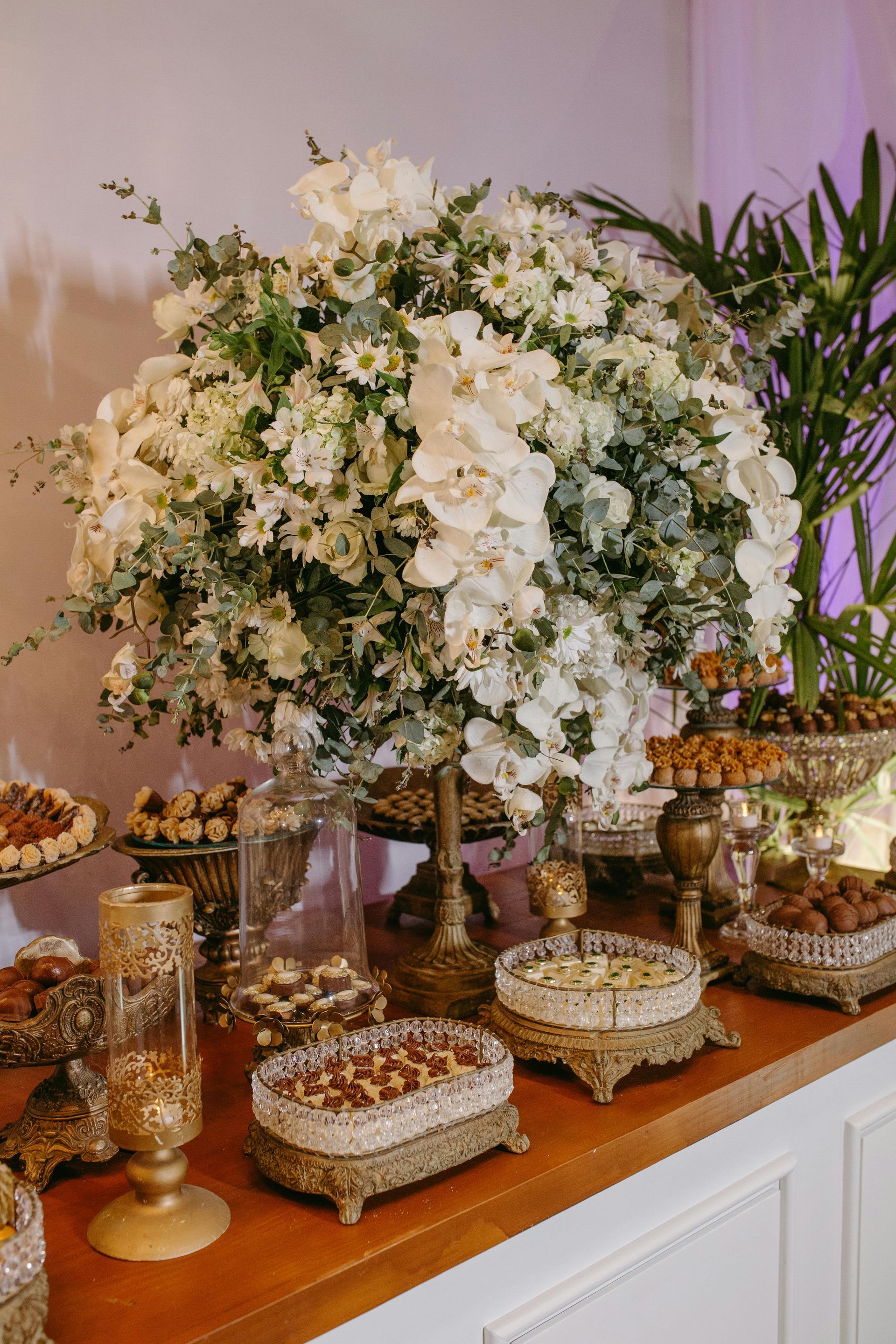 A dessert table with a large white floral arrangement. Sweets are displayed on decorative trays.